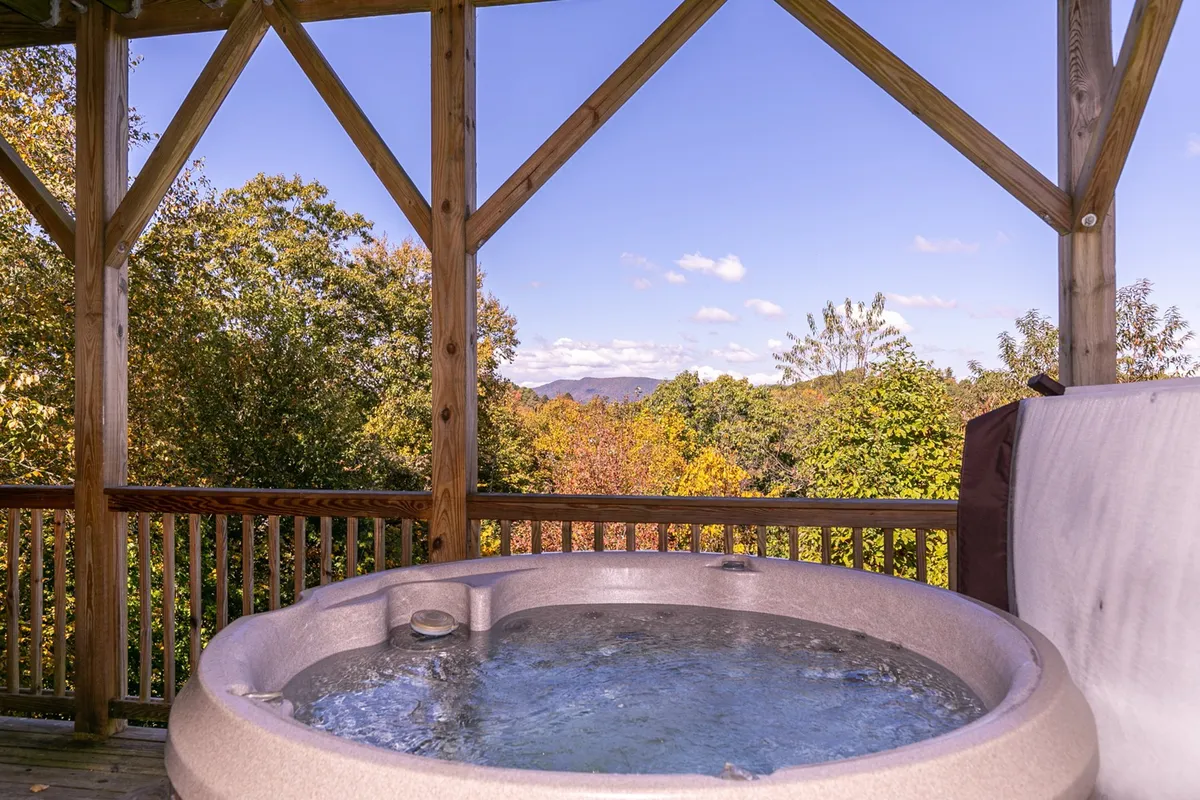 A spacious wooden deck featuring a hot tub, surrounded by lush trees, with expansive mountain views in the distance. The tranquil setting invites relaxation and stargazing, capturing the essence of soaking in nature's beauty from an elevated vantage point.