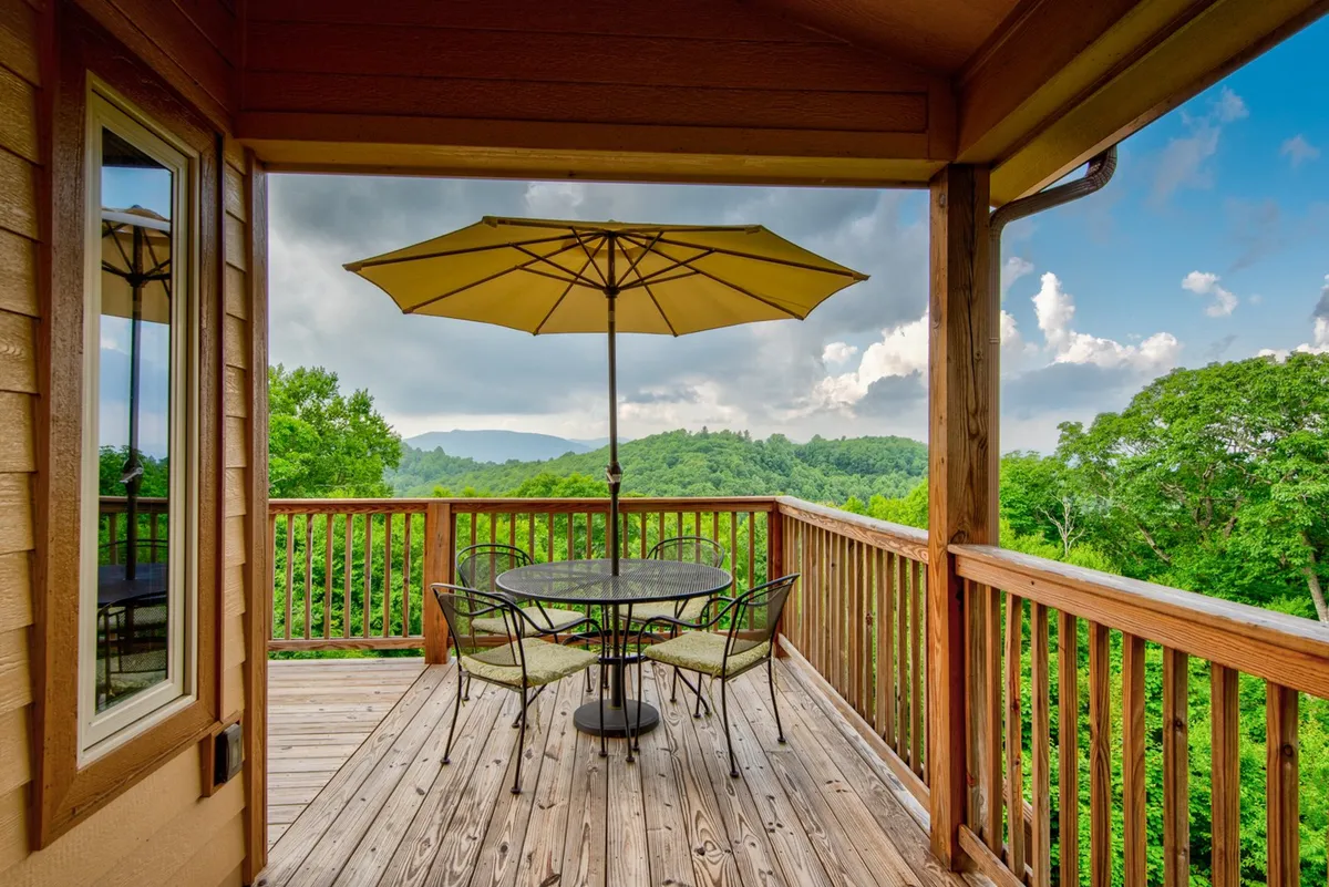 An exterior view of a deck with an outdoor dining table shaded by an umbrella, overlooking a scenic mountain landscape. The space is set for relaxation, morning coffee, or evening gatherings under the stars.