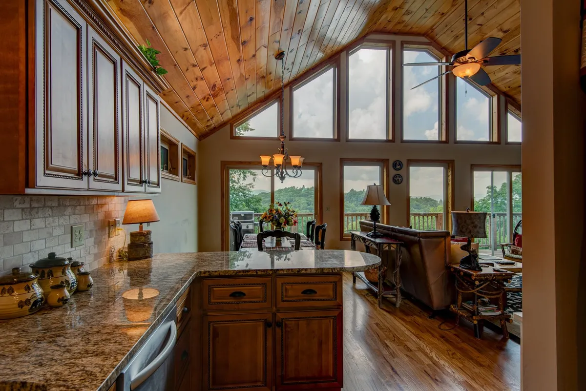 A spacious, light-filled open area that combines the dining table, kitchen with granite countertops, and large windows overlooking mountain ridges. The wooden ceiling and chandelier above the dining table create a warm, inviting atmosphere perfect for gatherings.