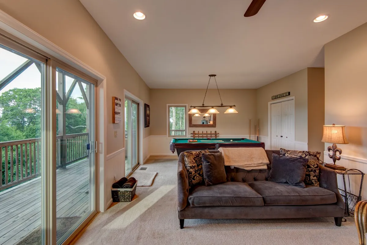 An interior recreation room with a pool table, comfortable sofa, and large windows that lead out to the deck, filled with natural light. The ambiance is casual and welcoming, ideal for friendly competitions and leisurely afternoons.