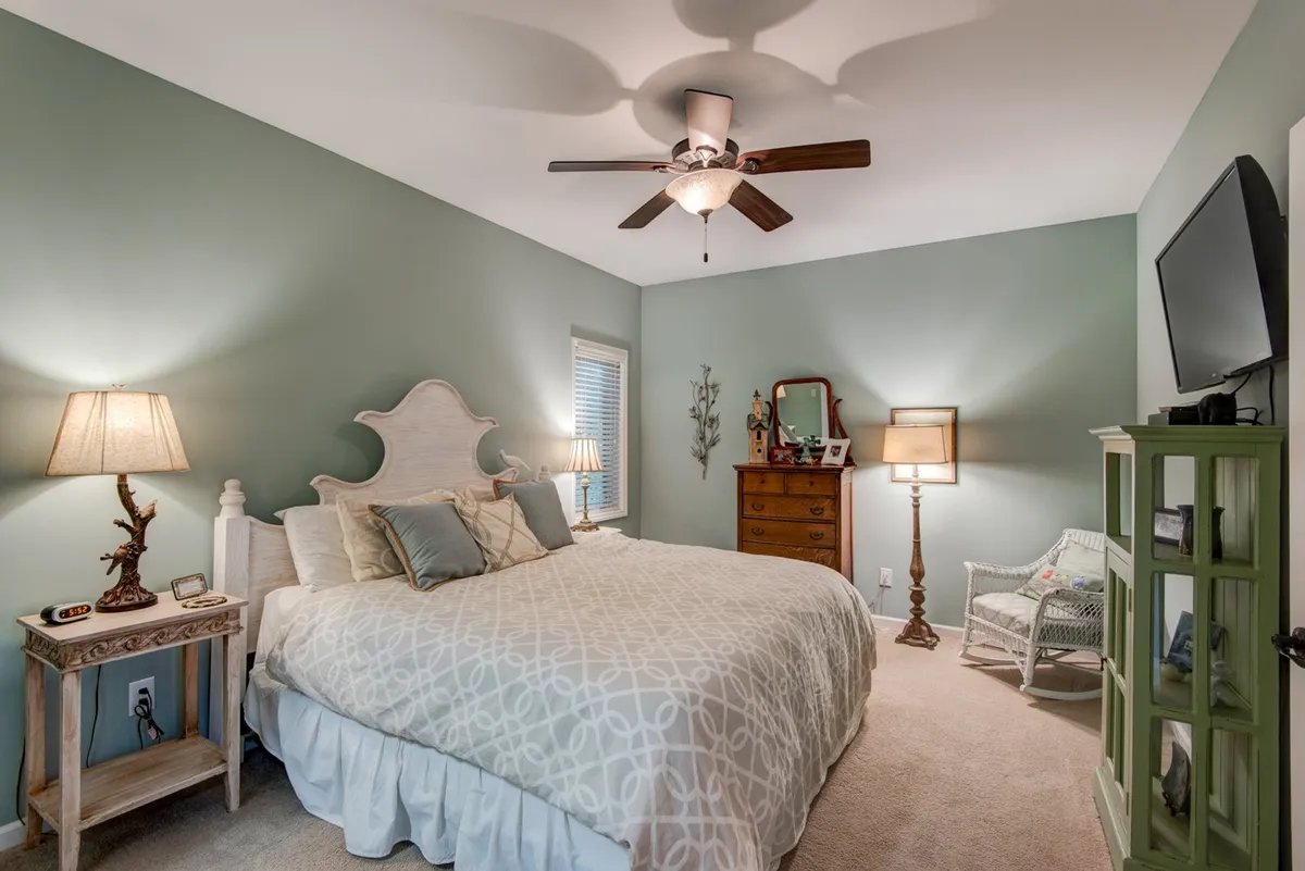 A serene bedroom featuring a king-sized bed with a decorative headboard and plush bedding, complemented by a nightstand with a lamp, a dresser with a mirror, green accent cabinet, and a cozy armchair. Gentle lighting and soft green tones create a tranquil and restful atmosphere.