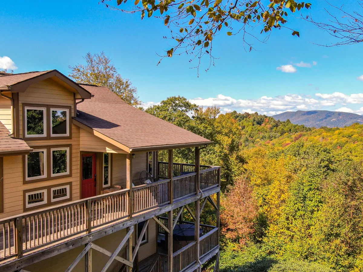 The image showcases a charming single-family home perched above rolling hills, its spacious wooden deck inviting guests to unwind. Surrounding the house, vibrant autumn foliage frames sweeping, unobstructed views of the Blue Ridge Mountains, with golden morning light hinting at tranquil sunrise moments. The scene is peaceful, evoking the serene mountain atmosphere unique to Blowing Rock.