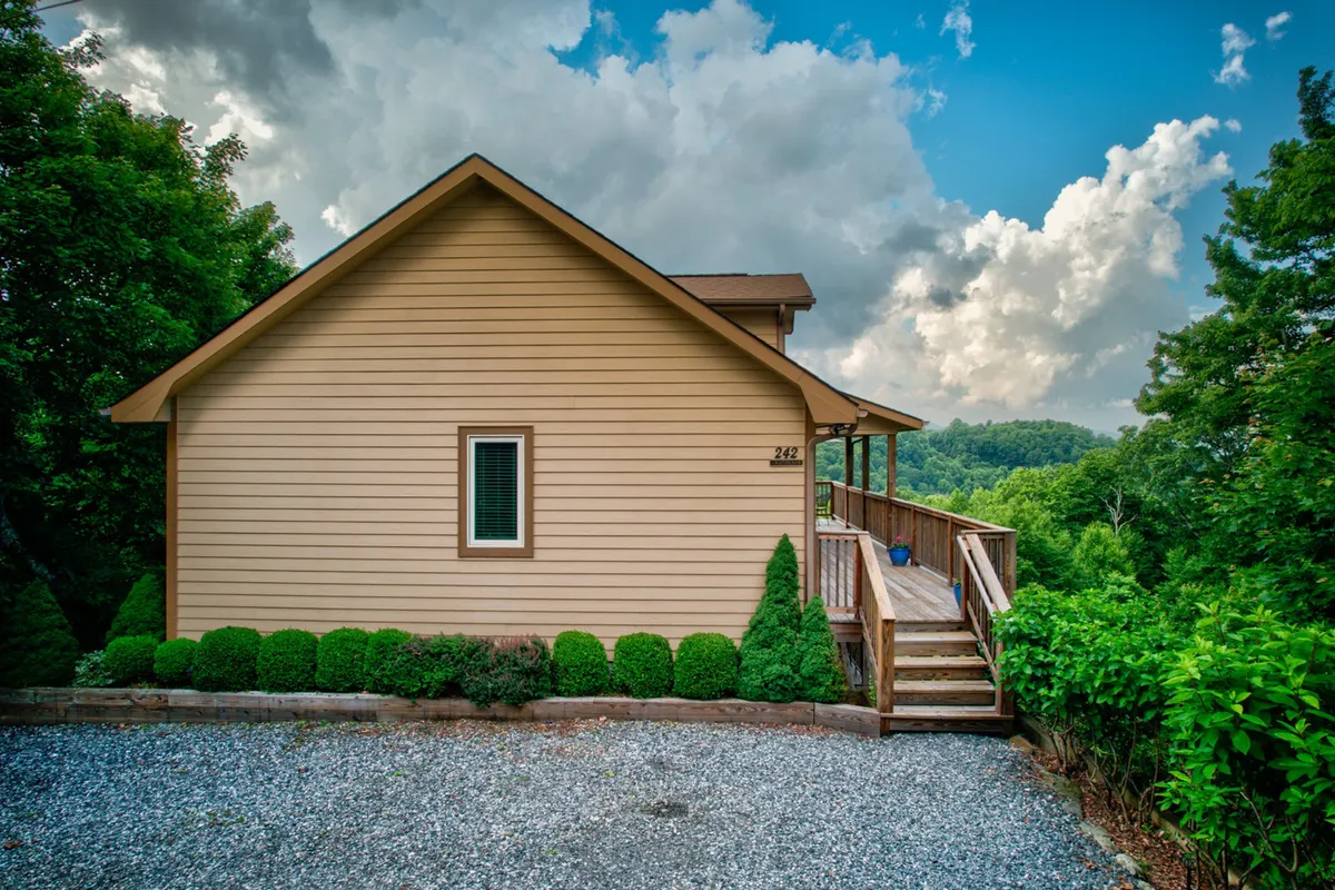 This image highlights the exterior of the home, including the wooden deck, landscaped garden, and large windows. The peaceful setting invites guests to imagine morning coffee or evening relaxation surrounded by mountain scenery.