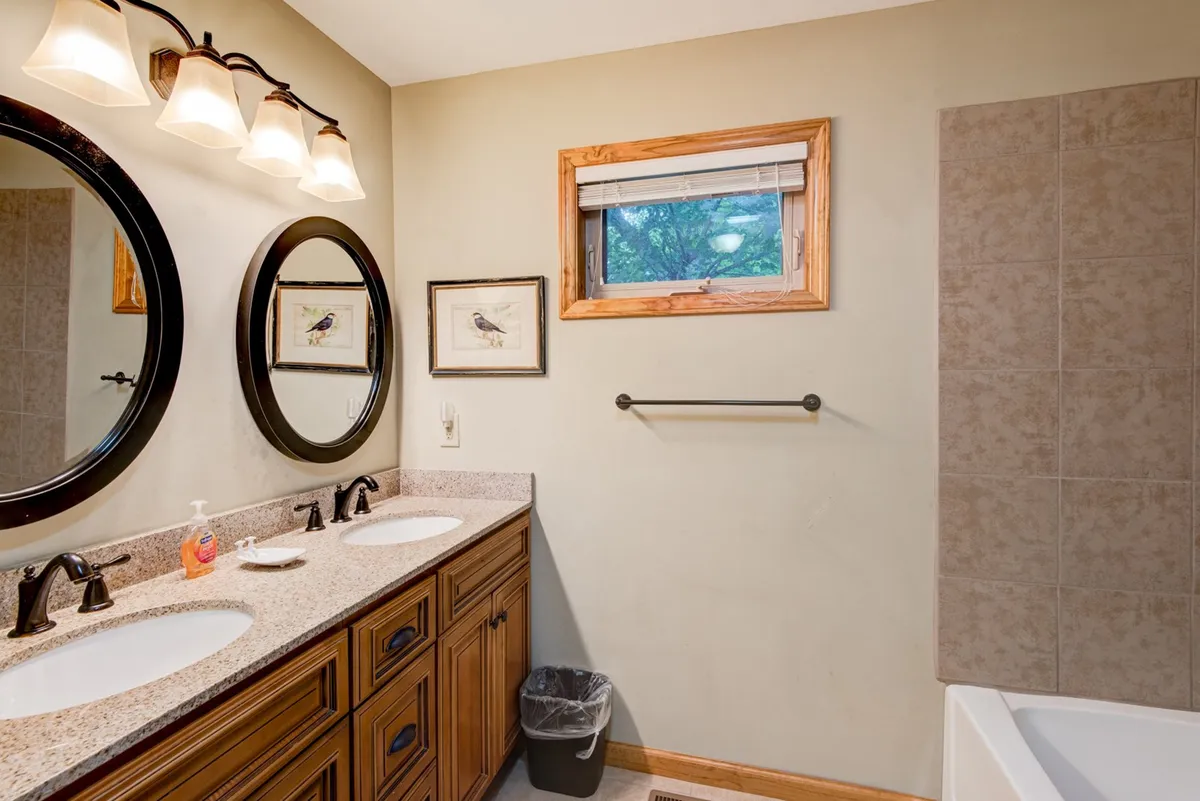 A spa-inspired en suite bathroom featuring a double vanity with two sinks, round mirrors, wall artwork, a window that brings in natural light, and a bathtub in the corner. The space exudes calm with neutral tones and a serene atmosphere ideal for self-care.