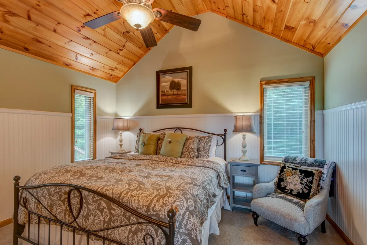 A tranquil bedroom with a bed featuring decorative bedding, an inviting armchair by the window, framed artwork, and stylish table lamps. Soft natural light streams in, creating a cozy and relaxing space perfect for unwinding or reading before sleep.