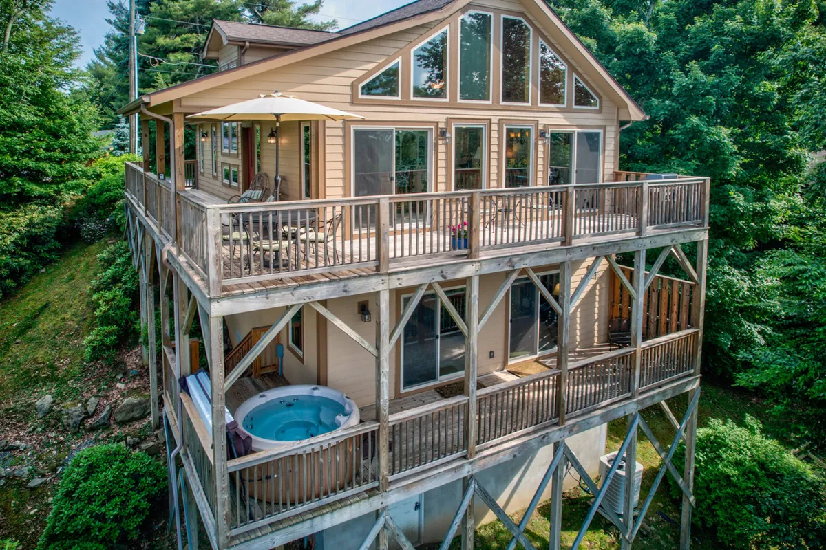 A wide view of the home's outdoor deck, featuring large windows, a hot tub, and ample deck space surrounded by lush greenery. The image captures the tranquil atmosphere with comfortable seating and sunset views over the mountains, emphasizing both relaxation and the natural setting.