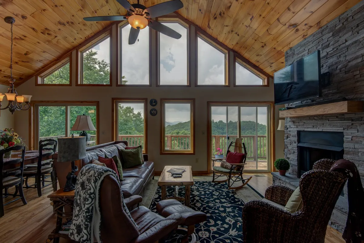 This image captures a comfortable great room with large windows framing sweeping mountain views. The stone fireplace anchors the space, complemented by a wooden ceiling and plush seating. The overall atmosphere is serene and welcoming, inviting relaxation while taking in the scenic landscape.