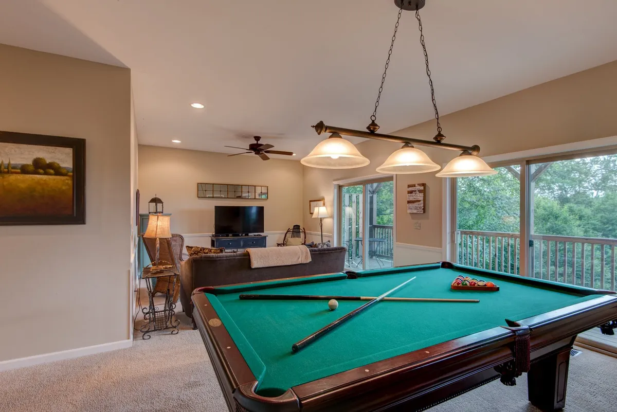 This cozy, entertainment-focused room displays both a pool table and a comfortable sofa facing windows, with a television visible in the frame. The layout encourages both socializing and movie nights.