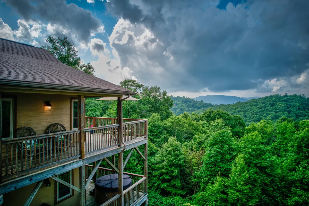 A wooden balcony overlooks a lush green landscape with a sweeping mountain range in the distance and a cloudy sky above. The scene suggests a tranquil mountain setting, perfect for admiring panoramic winter views when the landscape is dusted with snow.