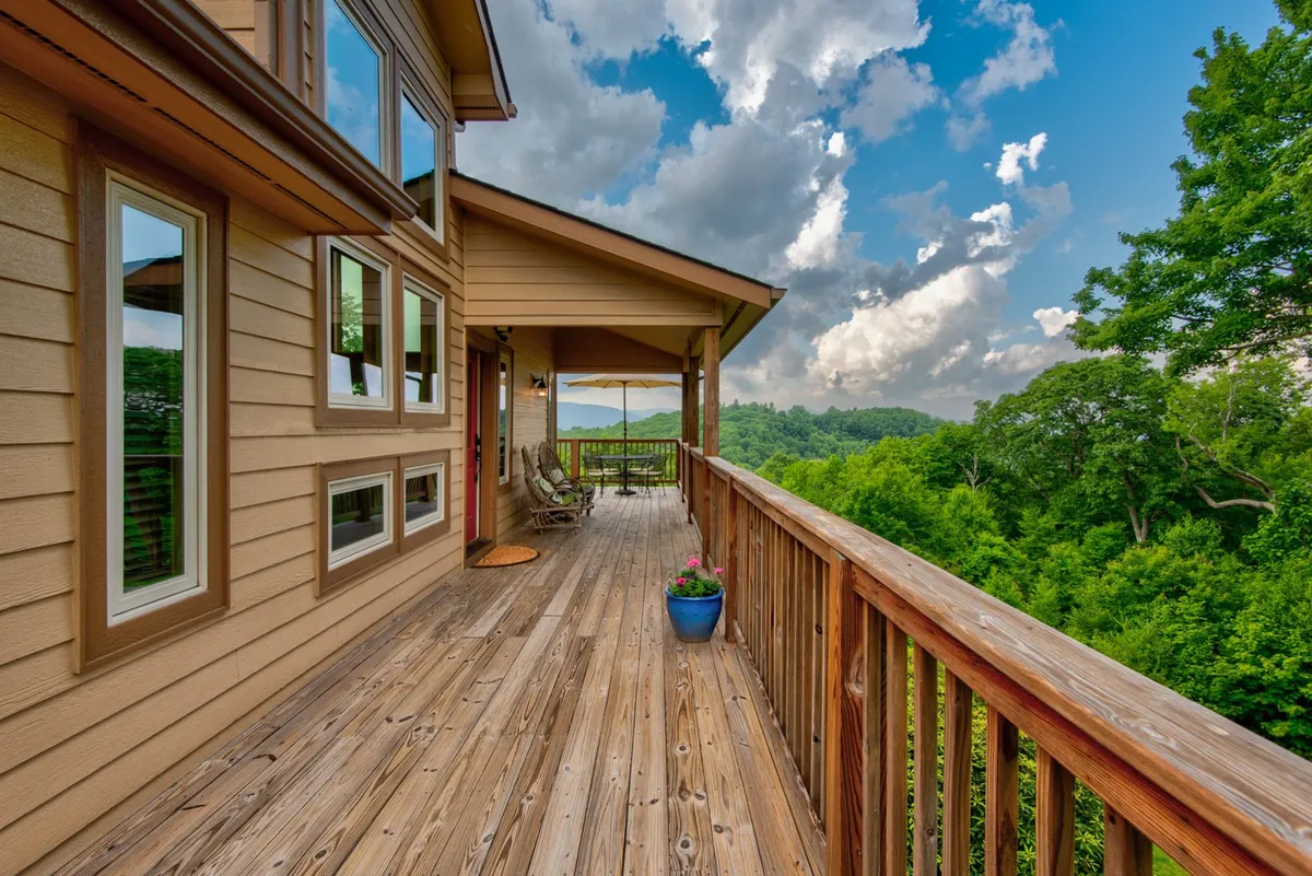 A peaceful wooden deck with rocking chairs, a planter with flowers, surrounding greenery, and distant mountains visible. The scene evokes a classic Appalachian porch experience, ideal for unwinding and observing the changing landscape throughout the seasons.